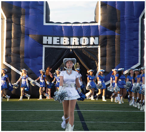 A person in a white uniform and hat stands on a football field holding pom-poms in front of a group of people in blue uniforms with pom-poms near a large inflatable structure with the word “HEBRON" on it. Description generated by AI