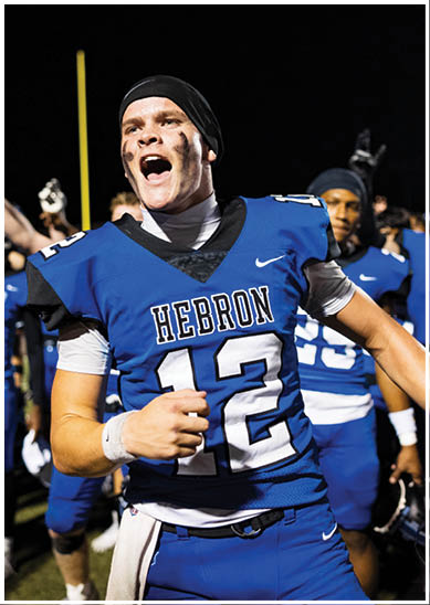 An adult wearing a blue Hebron football uniform with the number 12 is celebrating energetically on a field at night with teammates in the background. Description generated by AI