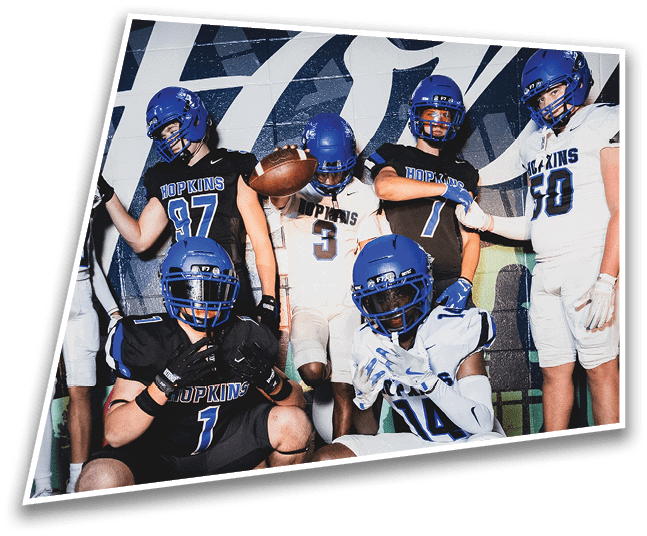 Four people in dark football uniforms and helmets stand in a row on a field, appearing focused and ready for action. Description generated by AI
