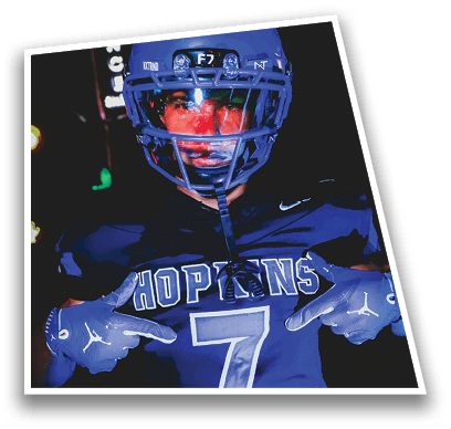 A person in football gear and helmet is crouched on a field with other players lined up in the background during a game. Description generated by AI