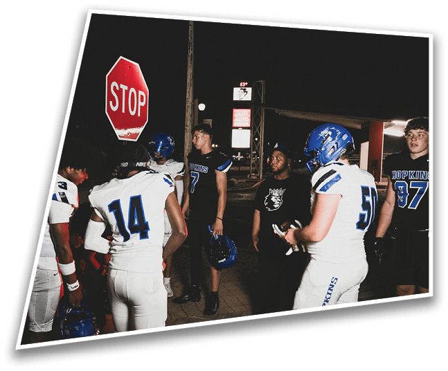 Four people in dark football uniforms and helmets stand in a row on a field, appearing focused and ready for action. Description generated by AI