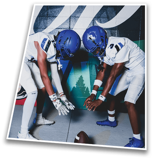 A person in a black football uniform and helmet is crouched in a ready position on a green field with another player in white uniform visible in the background. Description generated by AI