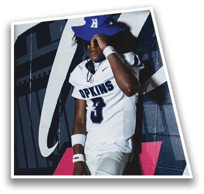 A person in football gear and helmet is crouched on a field with other players lined up in the background during a game. Description generated by AI