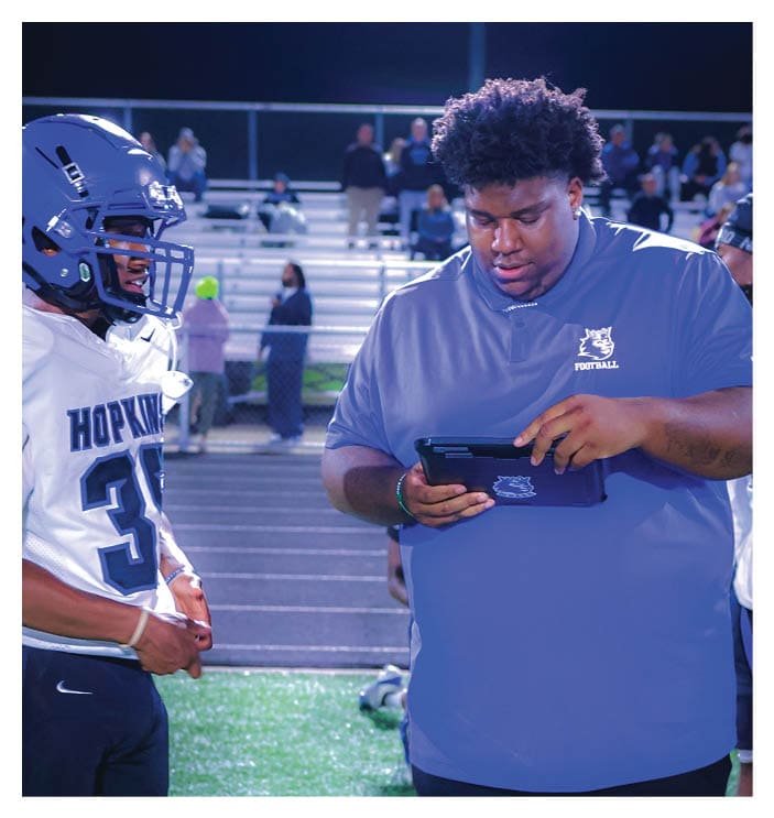 A football game is in progress with players on the field. One player is throwing a football while another player is trying to catch it. The players are wearing uniforms and are actively engaged in the game. AI generated content