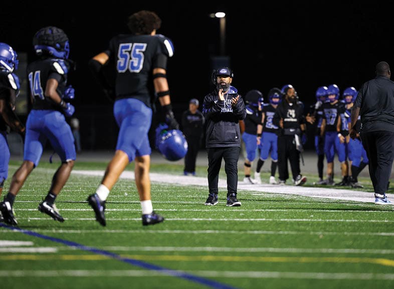 Four people wearing black football uniforms with “VISTA" on the front are standing together in a stadium with a crowd in the background, two of them holding helmets raised above their heads. Description generated by AI