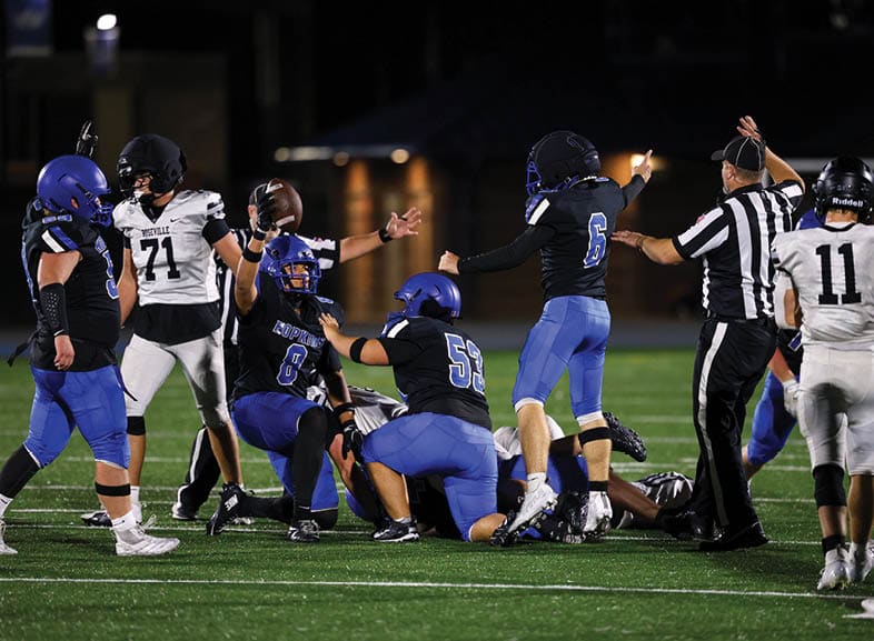 Four people wearing black football uniforms with “VISTA" on the front are standing together in a stadium with a crowd in the background, two of them holding helmets raised above their heads. Description generated by AI