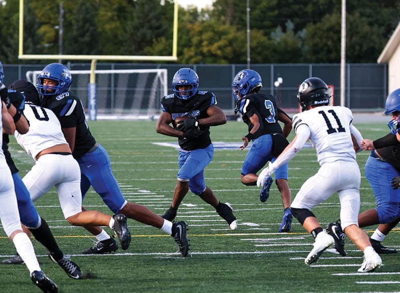 Four people wearing black football uniforms with “VISTA" on the front are standing together in a stadium with a crowd in the background, two of them holding helmets raised above their heads. Description generated by AI