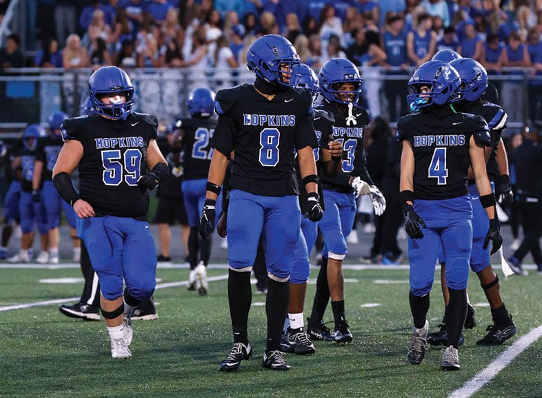 Four people wearing black football uniforms with “VISTA" on the front are standing together in a stadium with a crowd in the background, two of them holding helmets raised above their heads. Description generated by AI