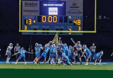 People in sports uniforms are running on a field with one person holding a football while others chase behind. Description generated by AI