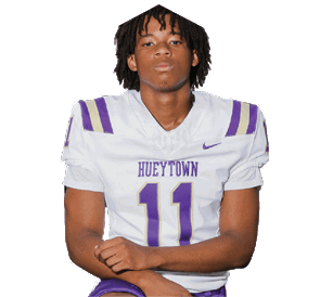 A person with dreadlocks wearing a white and purple football uniform kneels with one knee on the ground, holding a football, with two helmets placed in front. Description generated by AI