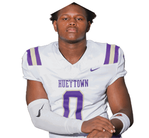 A person with short curly hair wearing a white football jersey with purple and gold stripes and the word “HUEYTOWN" on the front stands with arms crossed. Description generated by AI