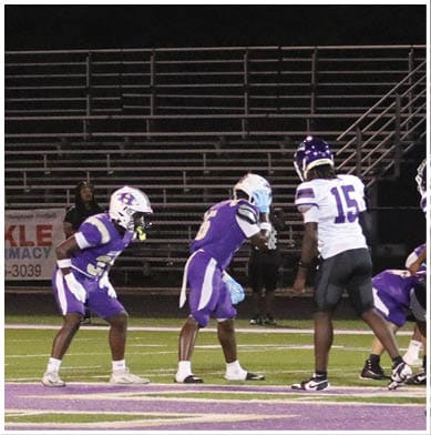 Four people in football uniforms and helmets are crouched in a ready stance on a field near a blue sideline. Description generated by AI