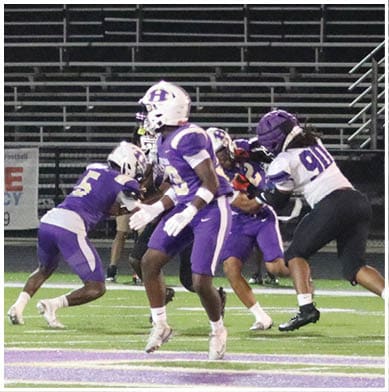 People in football uniforms are actively engaged in a play on a field with one person holding the ball and others positioned for blocking or defense. Description generated by AI