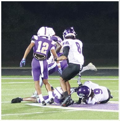 Four people in football uniforms and helmets are crouched in a ready position on a field near a blue sideline. Description generated by AI