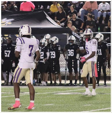 Four people in football uniforms and helmets are crouched in a ready stance on a field near a blue sideline. Description generated by AI