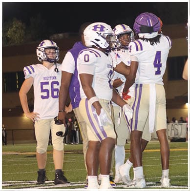 People in football uniforms are actively engaged in a play on a field with one person holding the ball and others positioned for blocking or defense. Description generated by AI