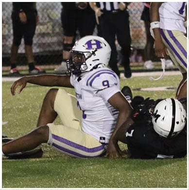 Four people in football uniforms and helmets are crouched in a ready position on a field near a blue sideline. Description generated by AI