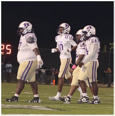 Four people in football uniforms and helmets are crouched in a ready position on a field near a blue sideline. Description generated by AI