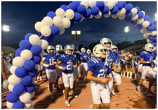Three football players in blue and white uniforms running on a field. Description generated by AI