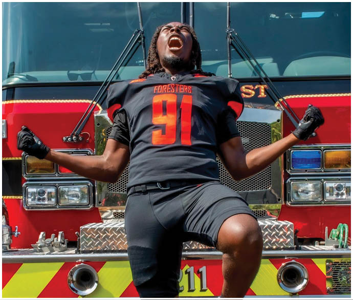 A man wearing a football jersey is standing in front of a fire truck. Description generated by AI