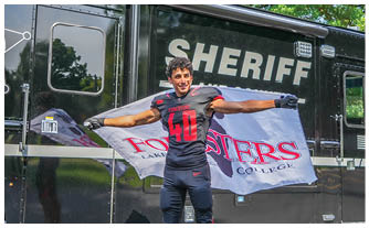 A man wearing a football jersey stands in front of a black truck with a sheriff logo on it. Description generated by AI