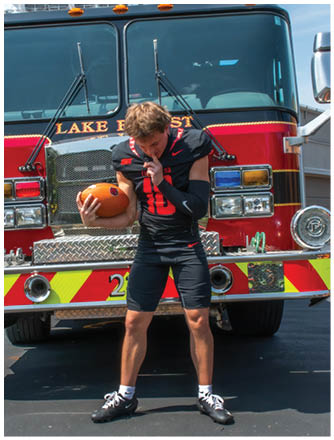 A man wearing a football jersey is holding a football in front of a fire truck. Description generated by AI