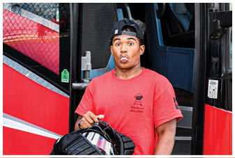 A man wearing a red shirt and black shorts is standing in front of a red and white bus. Description generated by AI
