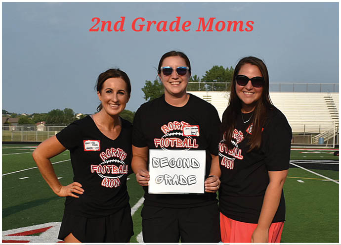 Three adults wearing matching black shirts with “North Football Moms" stand on a sports field with one holding a sign that says "Second Grade" under the red text "2nd Grade Moms." Description generated by AI