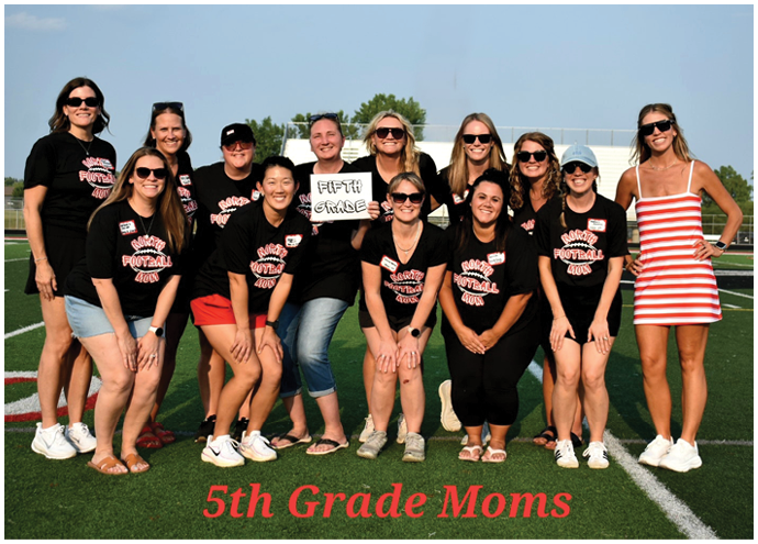 A group of adults wearing matching black shirts with pink text and one adult in a red and white striped dress are posing together on a sports field with a sign that reads “Fifth Grade" and the text "5th Grade Moms" displayed at the bottom. Description generated by AI