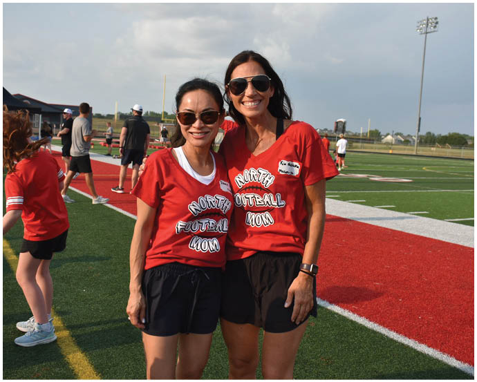 Two adults wearing matching red shirts with “Football Mom" text and sunglasses smile while standing on a sports field with other people in the background. Description generated by AI