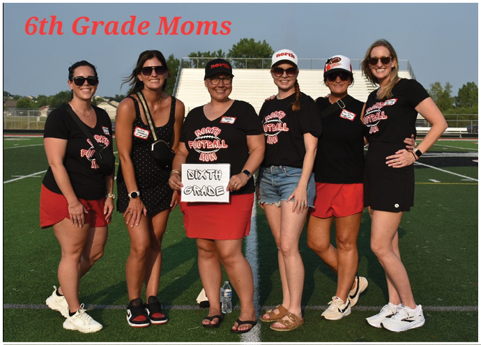 Six adults wearing matching sports-themed shirts and casual shorts or skirts stand together on a football field with one holding a sign that says “Sixth Grade." Description generated by AI
