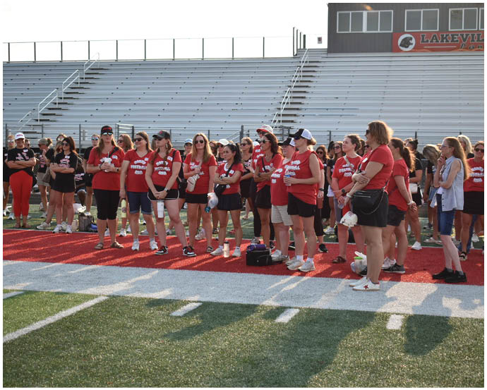 A group of people wearing matching red shirts and casual clothing stand together on a sports field near bleachers under a clear sky. Description generated by AI