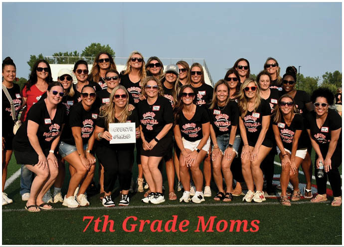 A group of adults wearing matching black shirts and sunglasses pose together on a sports field with a sign that reads “7th Grade Moms." Description generated by AI