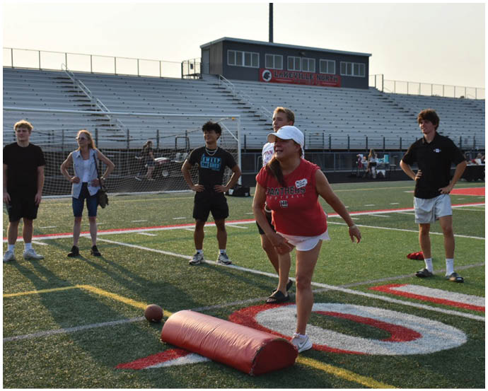 Several people wearing casual athletic clothing stand on a sports field with empty bleachers and a building labeled “Lakeville North" in the background. Description generated by AI