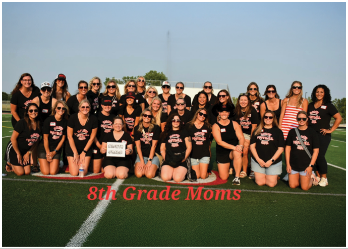 A large group of adults wearing matching black shirts and sunglasses pose together on a sports field with the text 8th Grade Moms displayed in front. Description generated by AI