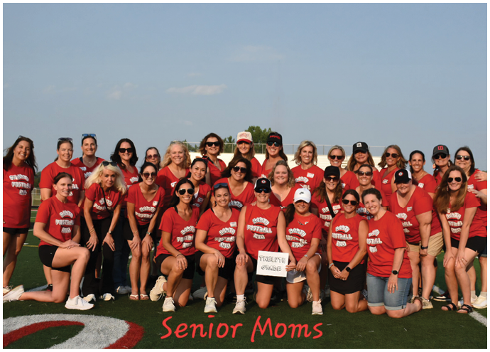 A group of adults wearing matching red shirts posing together on a sports field with the text “Senior Moms" at the bottom. Description generated by AI
