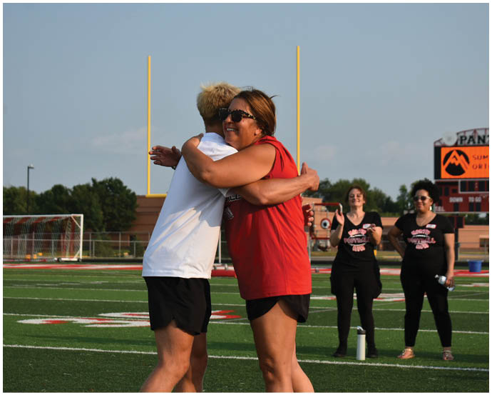 Two people are hugging on a sports field with three others standing in the background near a scoreboard that reads “Panthers." Description generated by AI