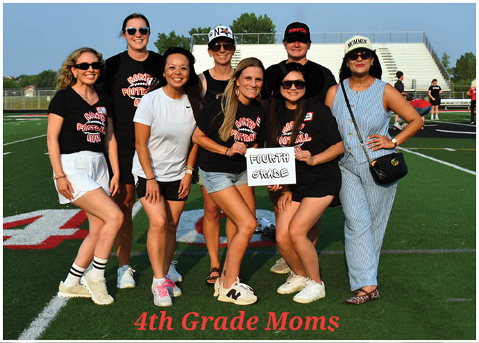 A group of eight adults smiling on a sports field with some wearing matching black shirts and one holding a sign that says “Fourth Grade." Description generated by AI
