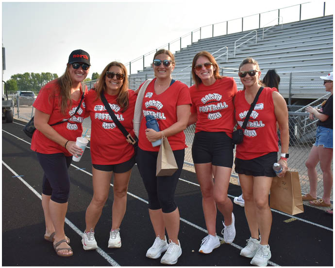 Five adults wearing matching red shirts with text and standing on a track field are smiling and posing for a photo in front of empty bleachers. Description generated by AI