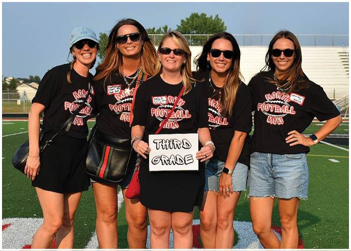 A group of eight adults smiling on a sports field with some wearing matching black shirts and one holding a sign that says “Fourth Grade." Description generated by AI