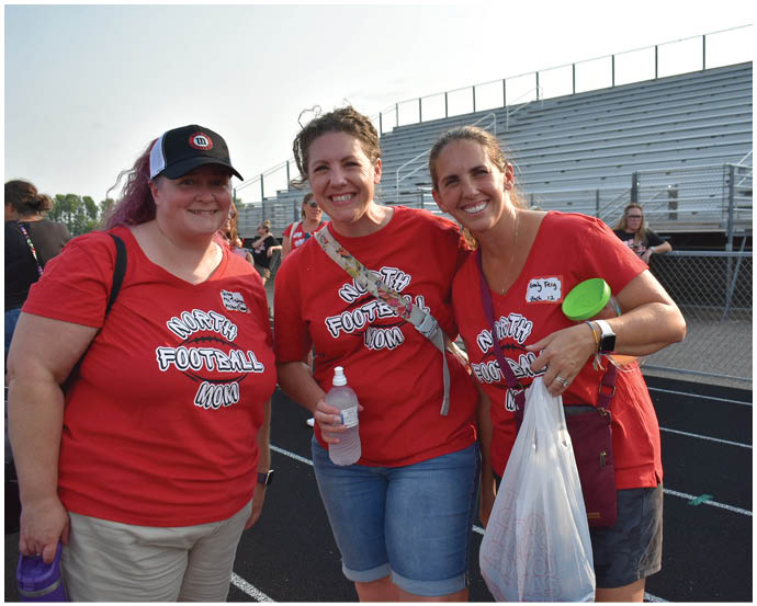 Three adults wearing matching red shirts that say “The Football Mom" are smiling and standing together at an outdoor sports venue with bleachers in the background. Description generated by AI