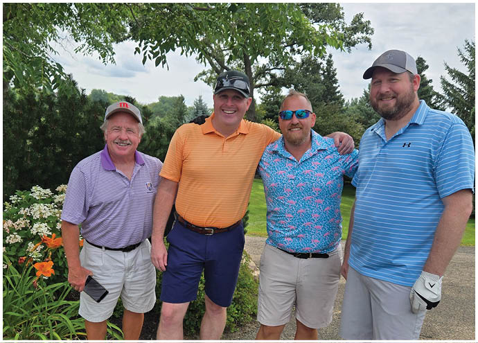 Four adults wearing casual golf attire stand closely together outdoors under a tree, smiling with arms around each other against a backdrop of greenery and a partly cloudy sky. Description generated by AI
