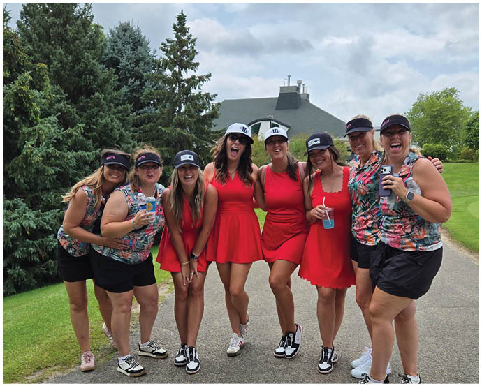 A group of eight people wearing colorful outfits and hats are standing closely together outdoors on a paved path with trees and a house in the background, smiling and holding drinks. Description generated by AI