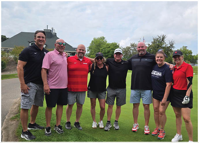 A group of eight adults dressed in casual and sporty clothing stand closely together on a golf course with trees and a building in the background. Description generated by AI