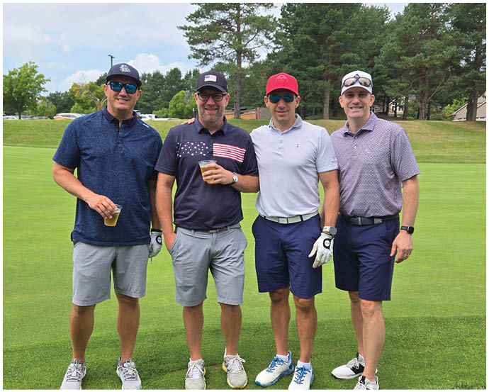 Four adults wearing casual golf attire and sunglasses stand together on a golf course with trees and a cloudy sky in the background. Description generated by AI