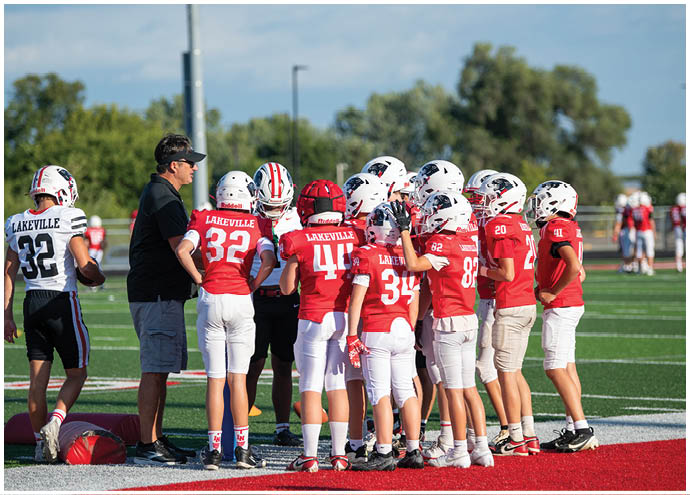A group of people in football uniforms and helmets are gathered on a field with one person in casual clothing appearing to give instructions. Description generated by AI
