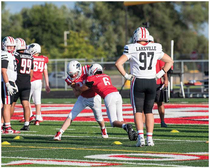 Adults wearing football uniforms are practicing on a field with one person tackling another while others stand nearby. Description generated by AI
