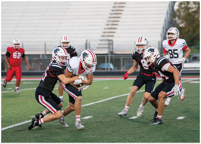 Several people wearing football uniforms are actively engaged in a tackle on a green field with empty bleachers in the background. Description generated by AI