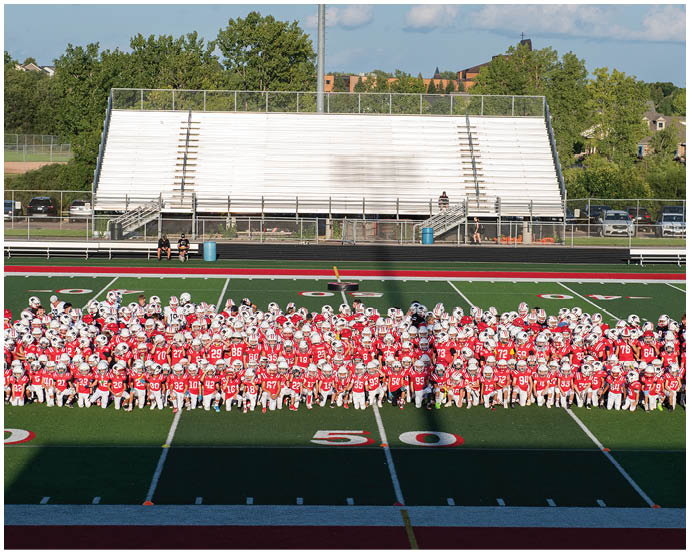A large group of people wearing red and white sports uniforms and helmets are gathered together on a football field in front of empty bleachers. Description generated by AI