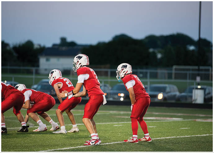 People in red and white football uniforms are lined up on a field preparing for a play during a game at dusk. Description generated by AI
